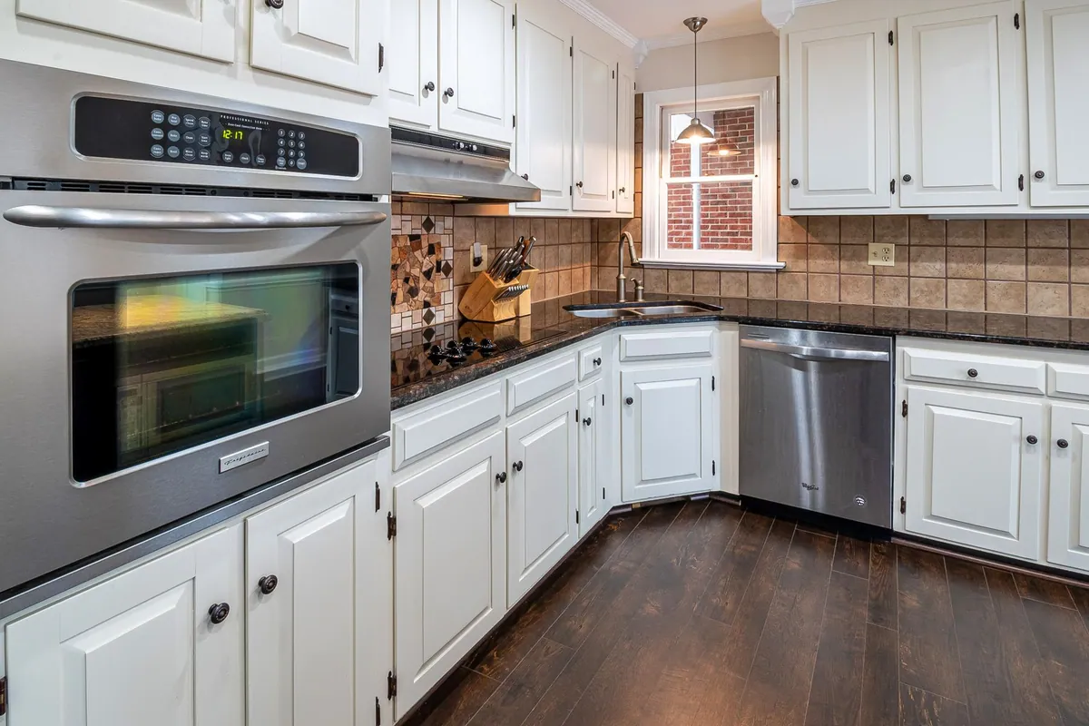 White painted kitchen cabinets with granite countertops, dark wood flooring, and stainless steel appliances